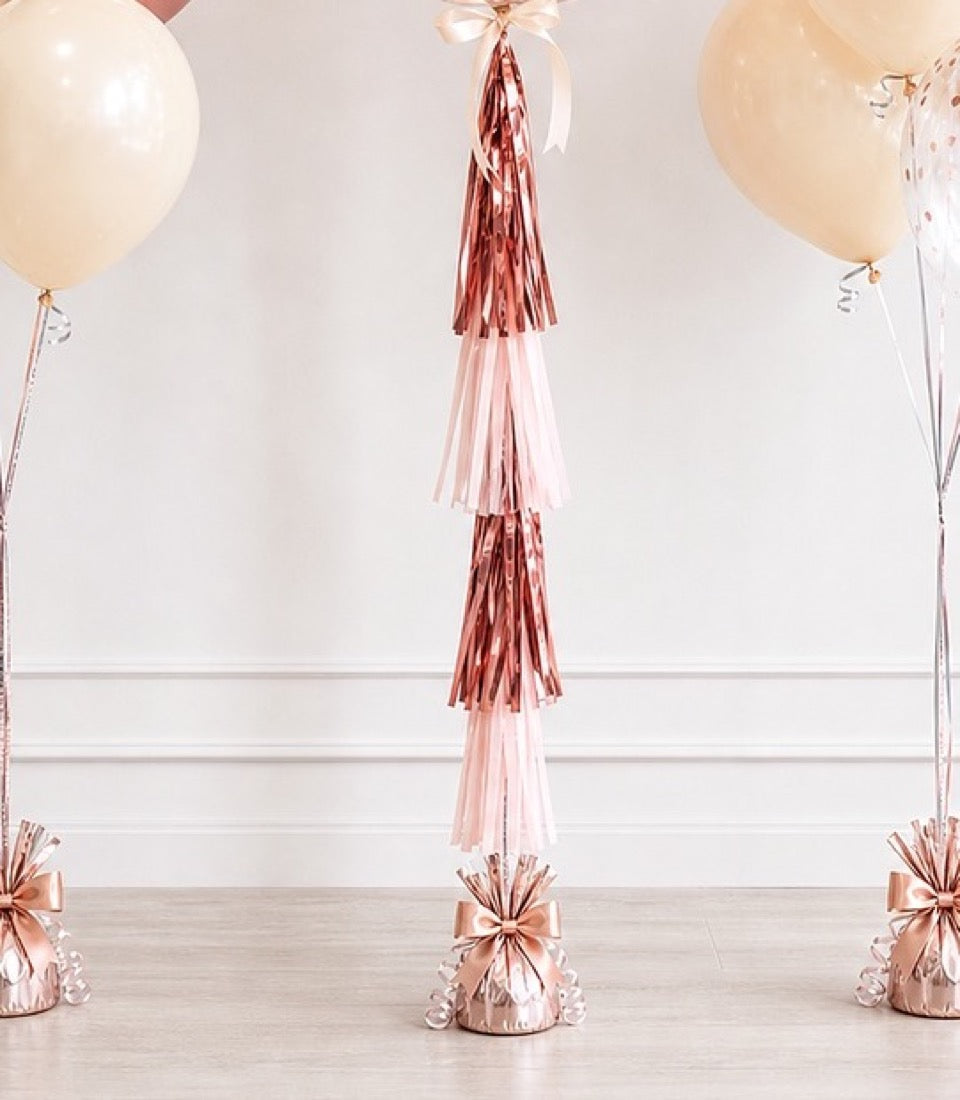 Rose gold tassel garland with alternating pink and burgundy metallic streamers, suspended between balloon weights on wooden floor.