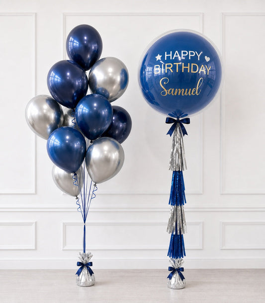 Navy blue and silver metallic balloons with personalized "Happy Birthday Samuel" bubble balloon, blue ribbons and tassels on white wall background.