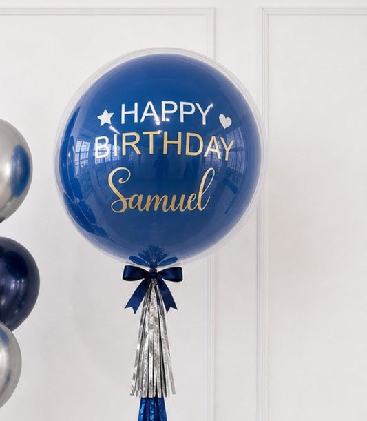 Blue bubble balloon with "Happy Birthday Samuel" text in white and gold, navy bow, and silver tassel streamer decoration.