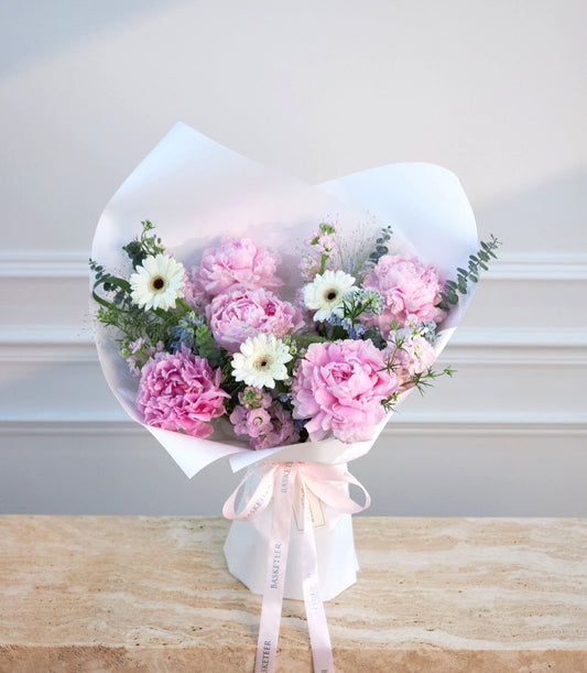 Elegant bouquet with pink peonies, white gerberas, and purple flowers wrapped in white paper with pink ribbon, displayed against a neutral background.
