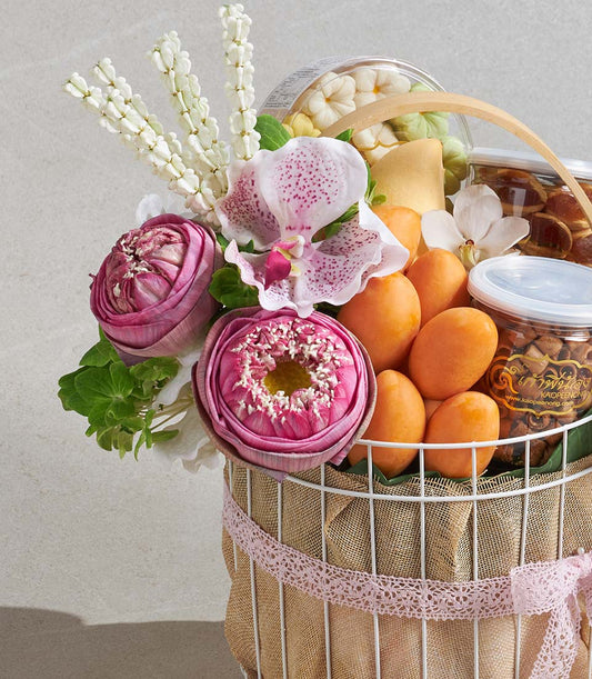 White wire gift basket with pink lotus flowers, fresh fruit, and gourmet treats in elegant pink and white arrangement