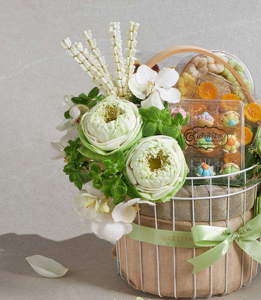 White wire gift basket with white lotus flowers, orchids, and Thai sweets in clear containers, tied with green Basketeer