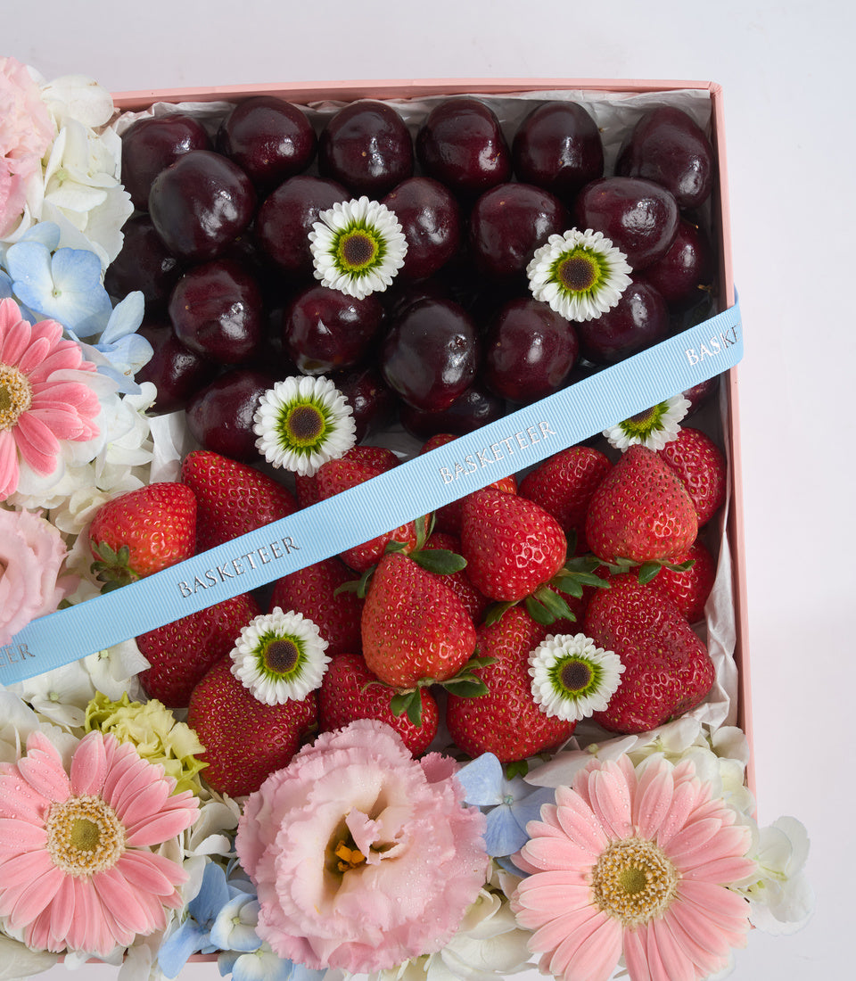 # Alt Text
Elegant gift box with dark cherries and red strawberries topped with white daisies, surrounded by pink and white flowers with blue ribbon.