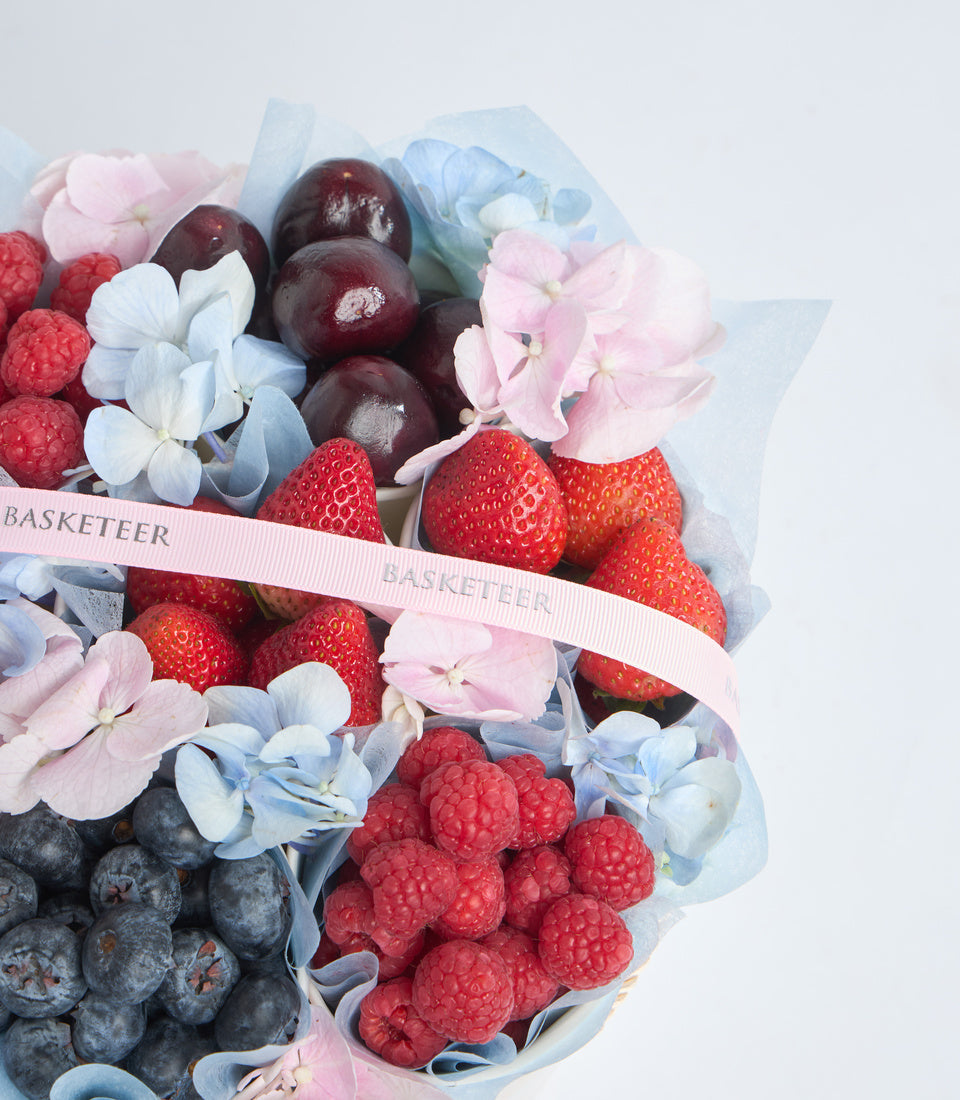 Luxury fruit and flower gift basket with strawberries, raspberries, blueberries, dark plums, pink hydrangeas, and blue flowers wrapped in tissue paper with pink ribbon.