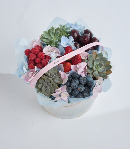 Luxury fruit and succulent arrangement in white ceramic bowl with light blue tissue, fresh berries, pink flowers, and pink ribbon labeled "Basketeer."