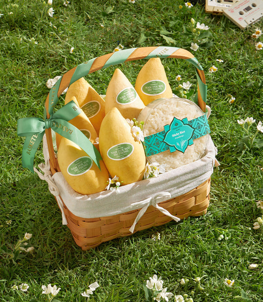Woven picnic basket filled with golden Nam Dok Mai mangoes and sticky rice, set on green grass with vintage aesthetic
