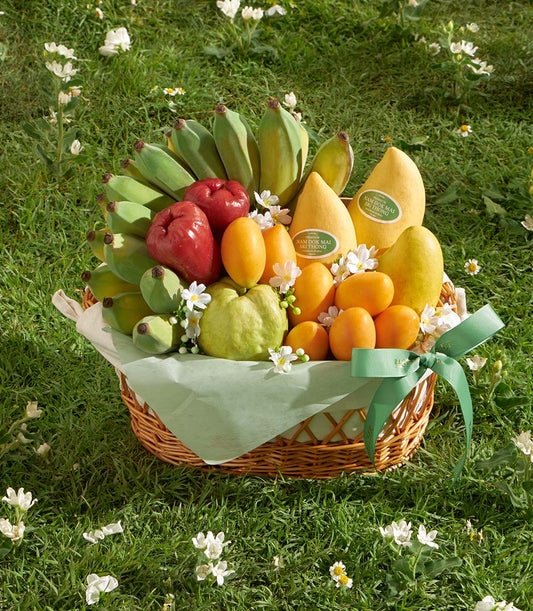 Wicker basket filled with fresh Thai mangoes, bananas, guavas, and citrus fruits on green tissue paper with white flowers