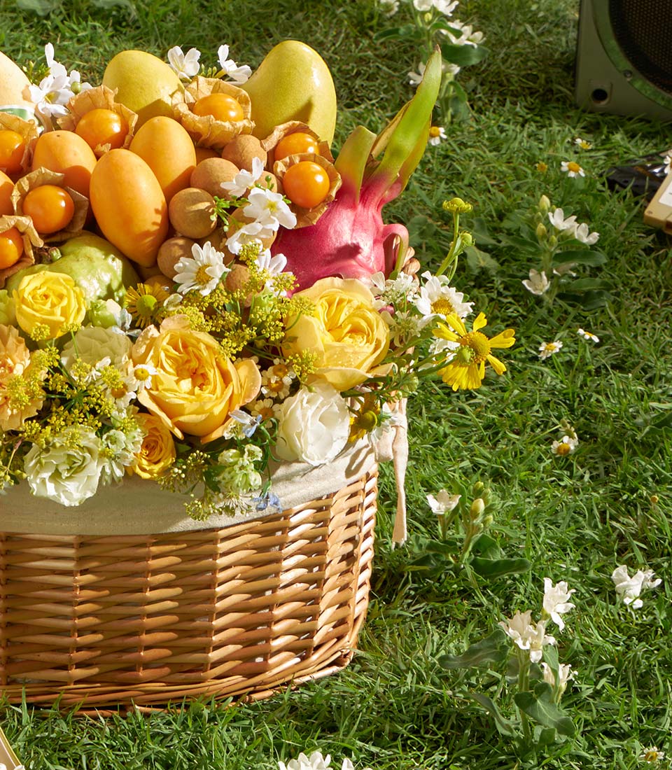 Wicker basket with tropical fruits, yellow roses, and daisies on green lawn