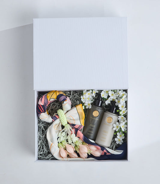 Luxury gift box containing colorful silk scarf, two dark bottles with gold seals, white daisy flowers, and green decorative filler on white background.