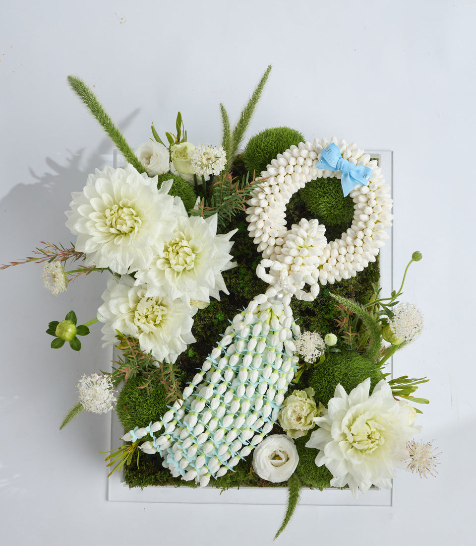 Luxurious floral arrangement featuring white dahlias, baby's breath wreath, moss balls, ornamental grasses, and blue ribbon bow on white square base.