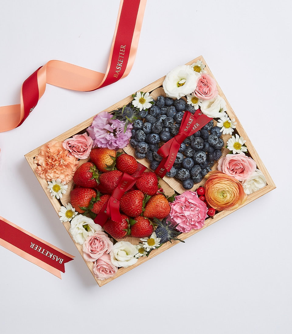 Wooden gift box with fresh red strawberries, blueberries, and mixed pink roses, carnations, daisies arranged with red ribbon