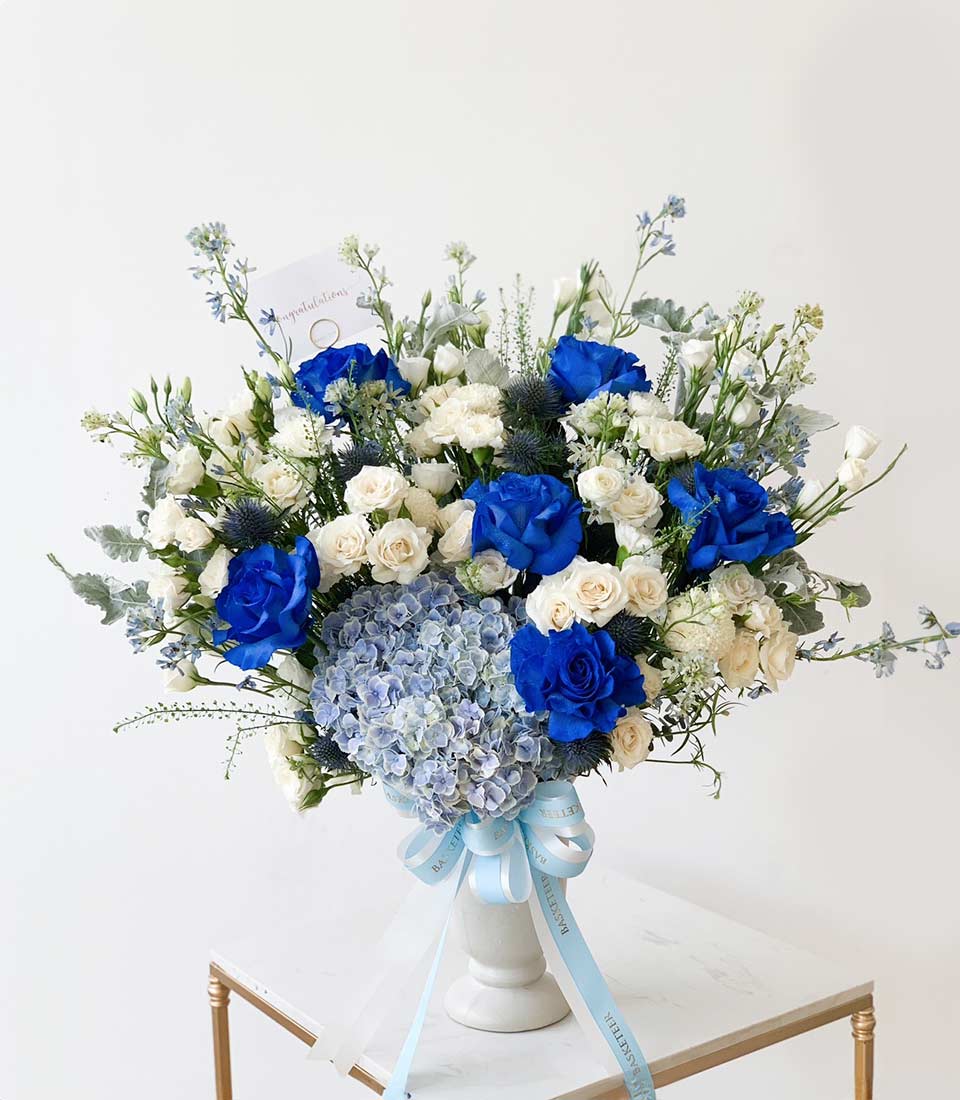 Elegant blue and white floral arrangement with roses, hydrangeas, and thistle in white vase with ribbon