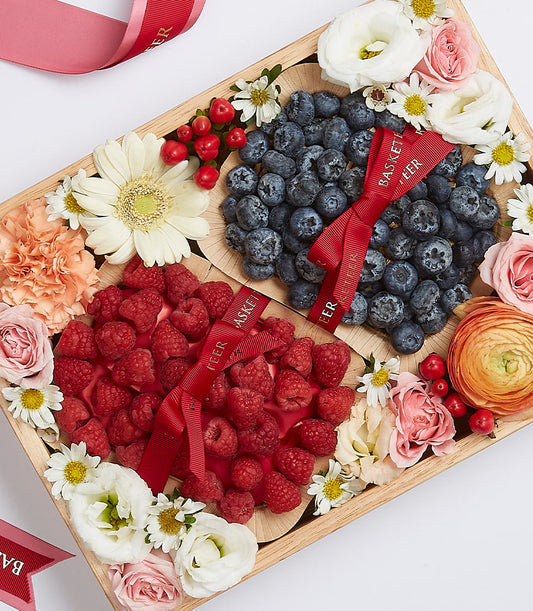 Wooden gift box with fresh raspberries and blueberries, surrounded by white daisies, pink roses, and orange ranunculus