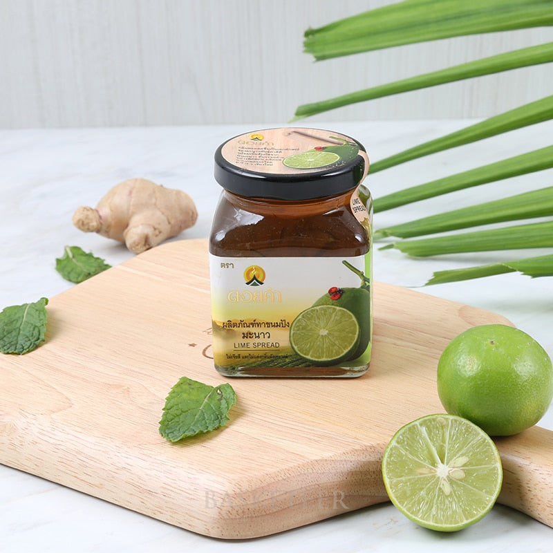 Lime spread in glass jar with black lid, surrounded by fresh ginger, mint leaves, and whole limes on wooden board