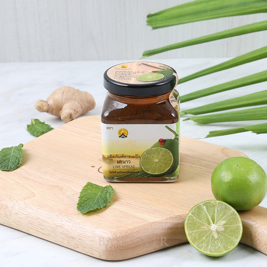 Lime spread in glass jar with black lid, surrounded by fresh ginger, mint leaves, and whole limes on wooden board