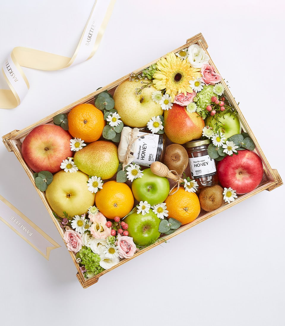 Wooden gift crate with fresh apples, oranges, pears, kiwis, mixed flowers, and honey jars