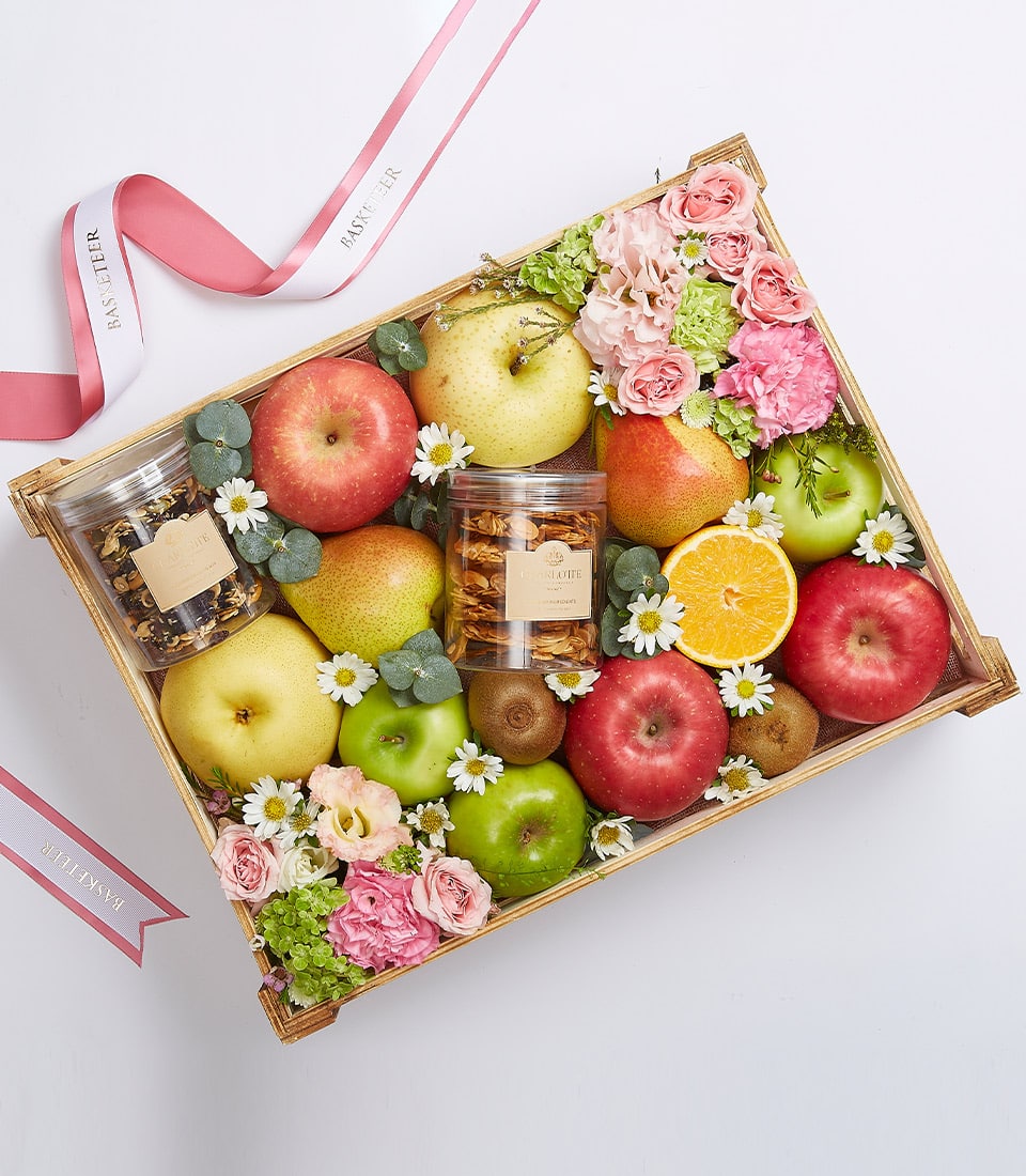 Wooden crate filled with fresh apples, pears, kiwis, orange slices, artisan snacks, and pink roses with eucalyptus
