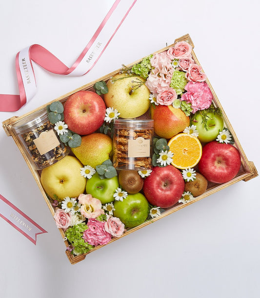 Wooden crate filled with fresh apples, pears, kiwis, orange slices, artisan snacks, and pink roses with eucalyptus