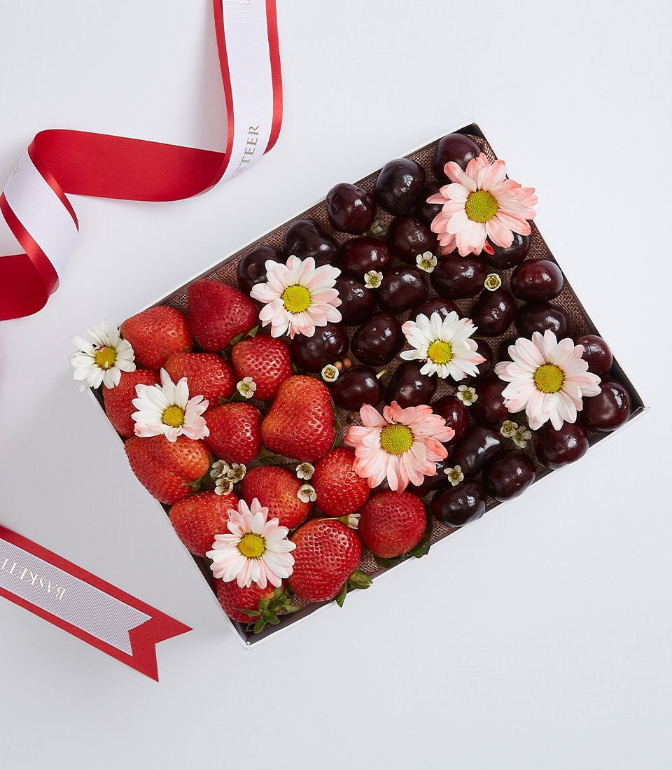 Rectangular gift box with fresh strawberries, dark cherries, white daisies, and delicate blossoms on burlap, tied with red