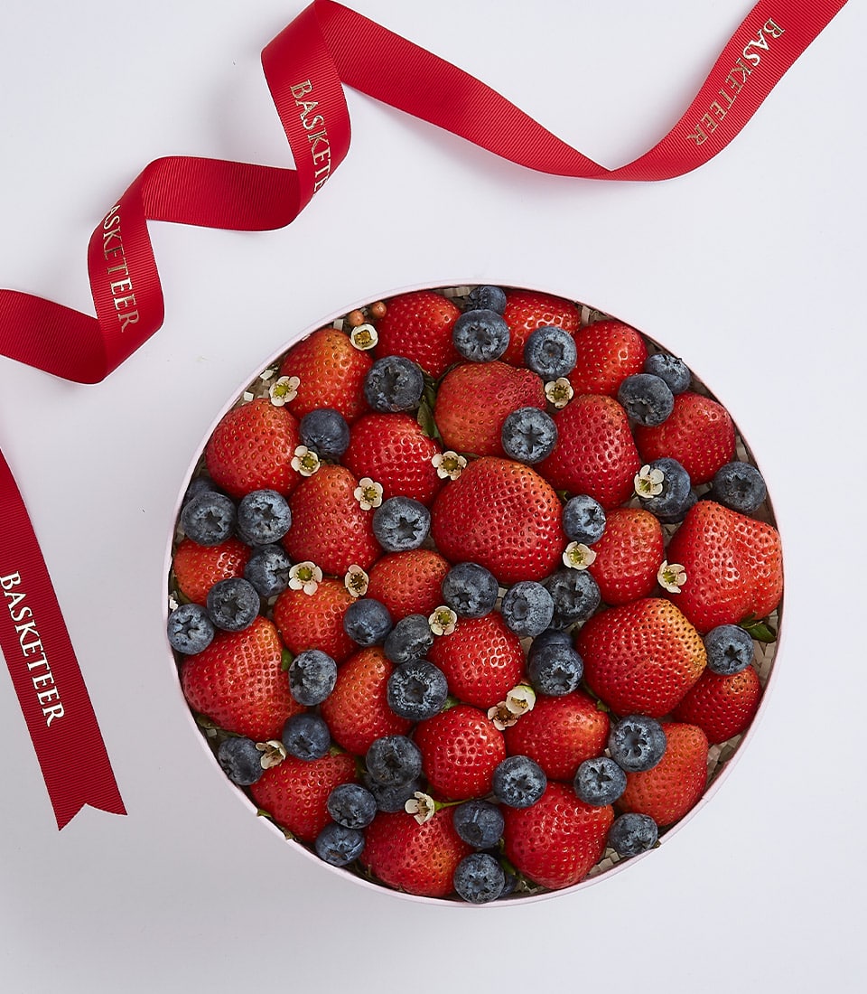 Light pink round gift box filled with fresh strawberries and blueberries with white flowers and gold-lettered red ribbon