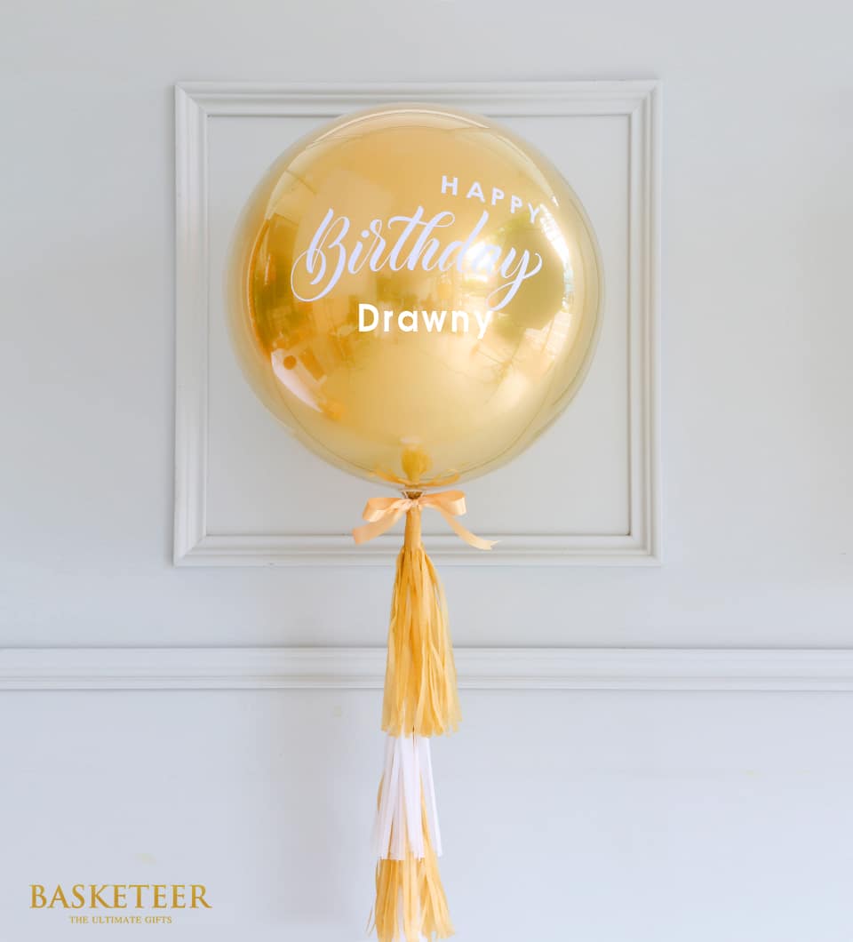 Golden helium balloon with personalized white text, peach satin ribbon, and golden tissue tassel garland for birthday gifting