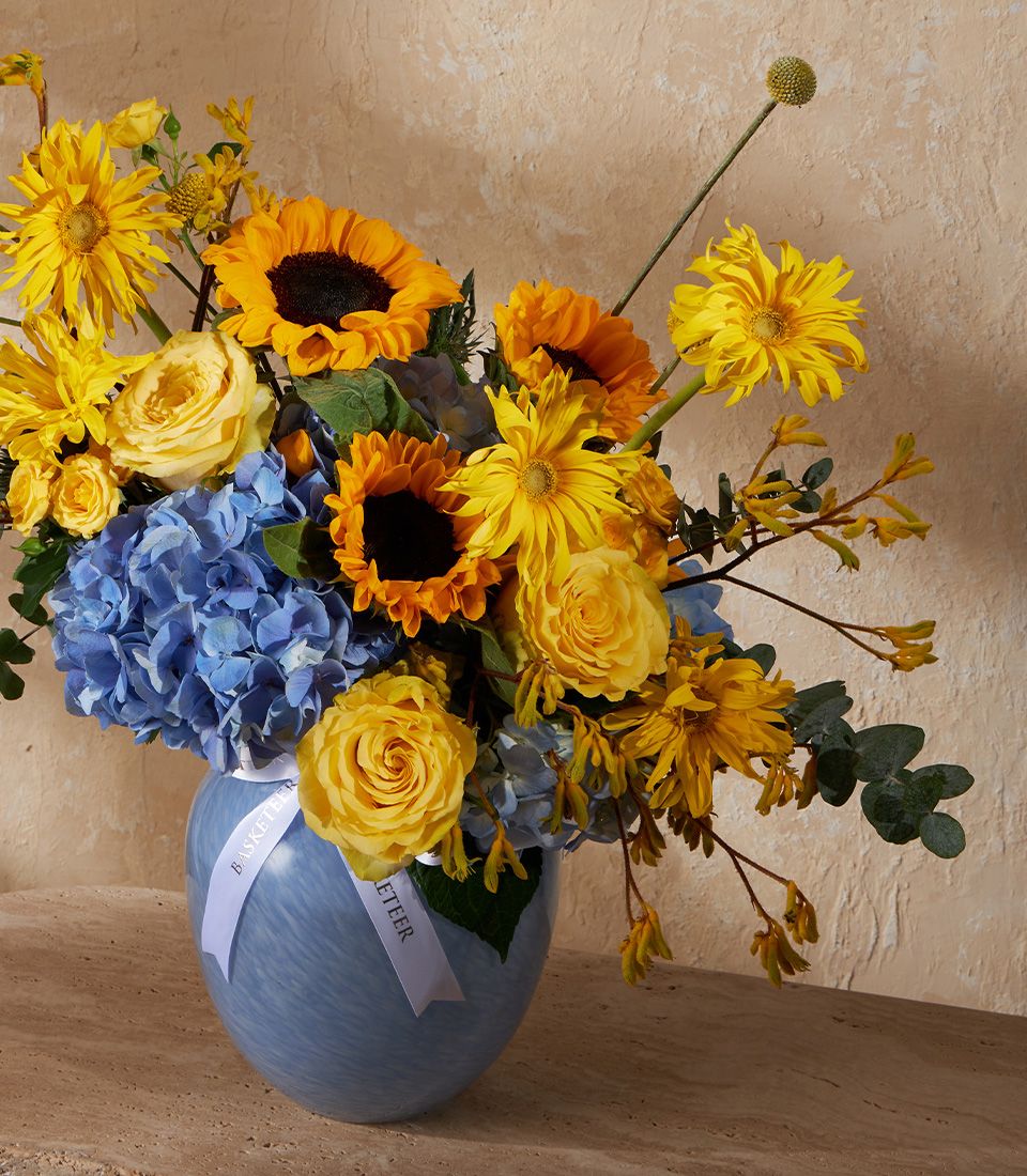 Vibrant yellow and blue floral arrangement in light blue swirled vase with sunflowers, roses, and hydrangeas