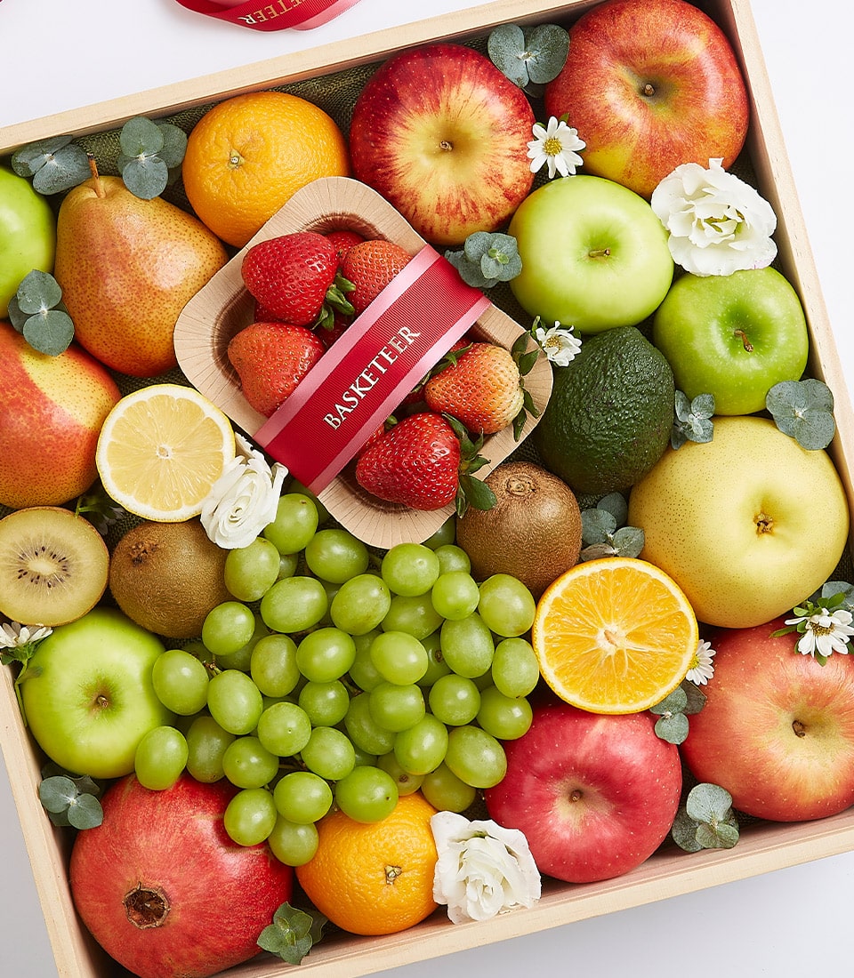 Wooden gift box of fresh fruits with apples, oranges, pears, kiwis, and strawberries in small bamboo bowl with red ribbon