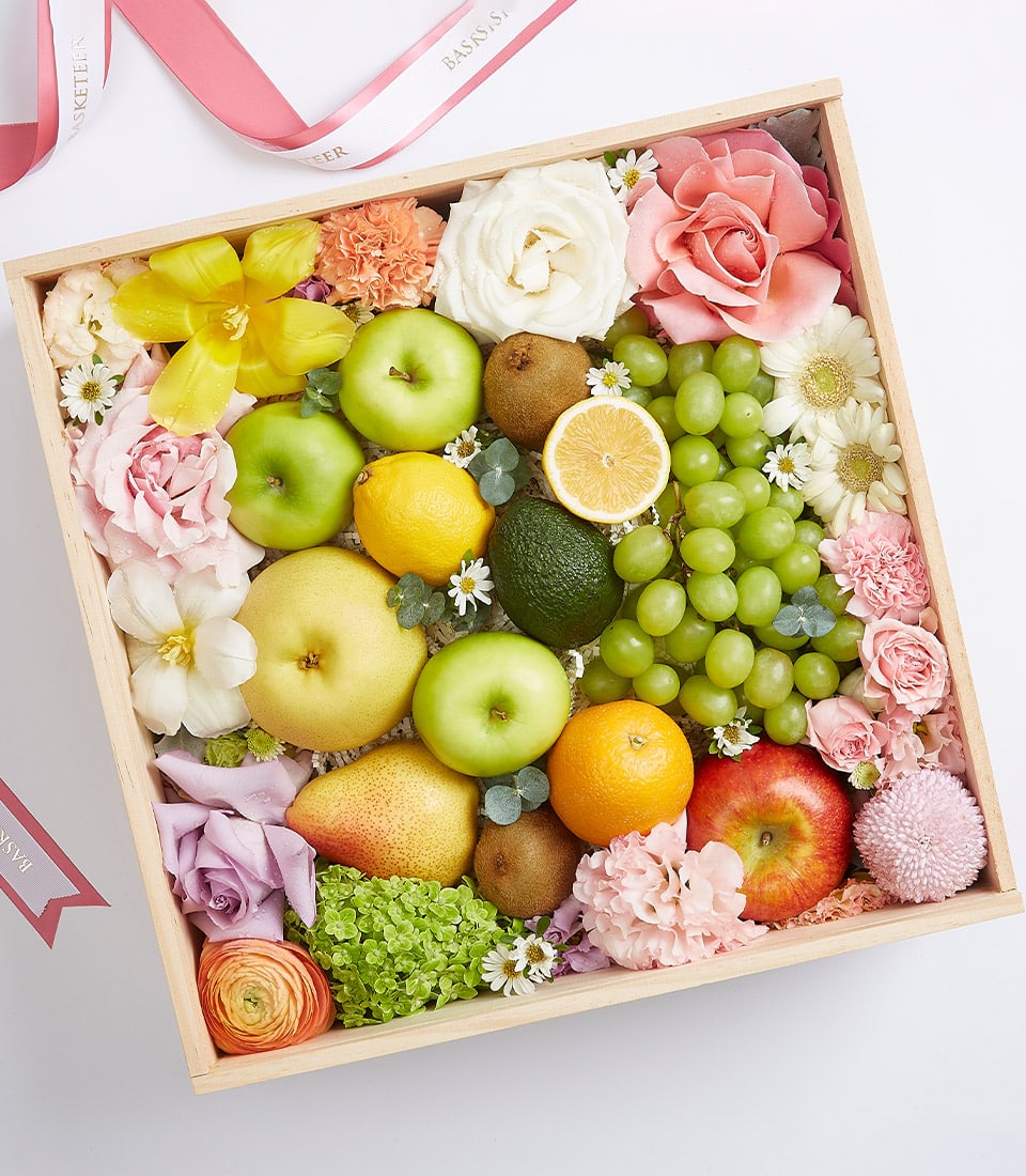 Wooden gift box with fresh apples, pears, citrus, kiwis and seasonal fruits, arranged with pink roses, tulips, and mixed