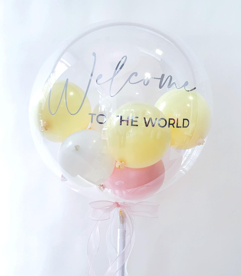 Clear bubble balloon with pale yellow, white, and pink balloons inside, "Welcome to the World" message, and light pink