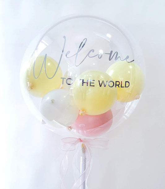 Clear bubble balloon with pale yellow, white, and pink balloons inside, "Welcome to the World" message, and light pink