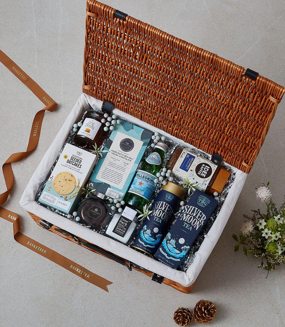 Light brown wicker picnic basket with gourmet tea, coffee, chocolate, oatcakes, jam and sparkling water, lined with white