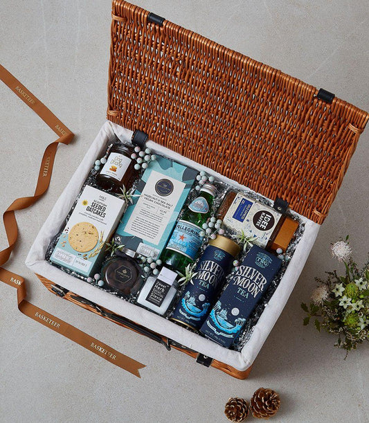 Light brown wicker picnic basket with gourmet tea, coffee, chocolate, oatcakes, jam and sparkling water, lined with white