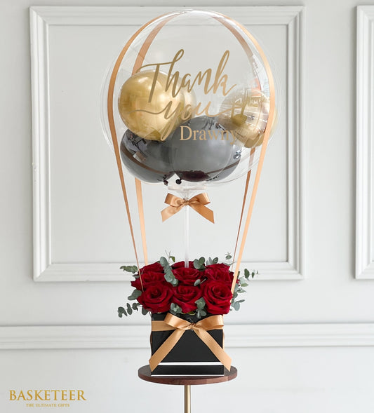 Black and gold hot air balloon arrangement with red roses and eucalyptus in suspended gift box