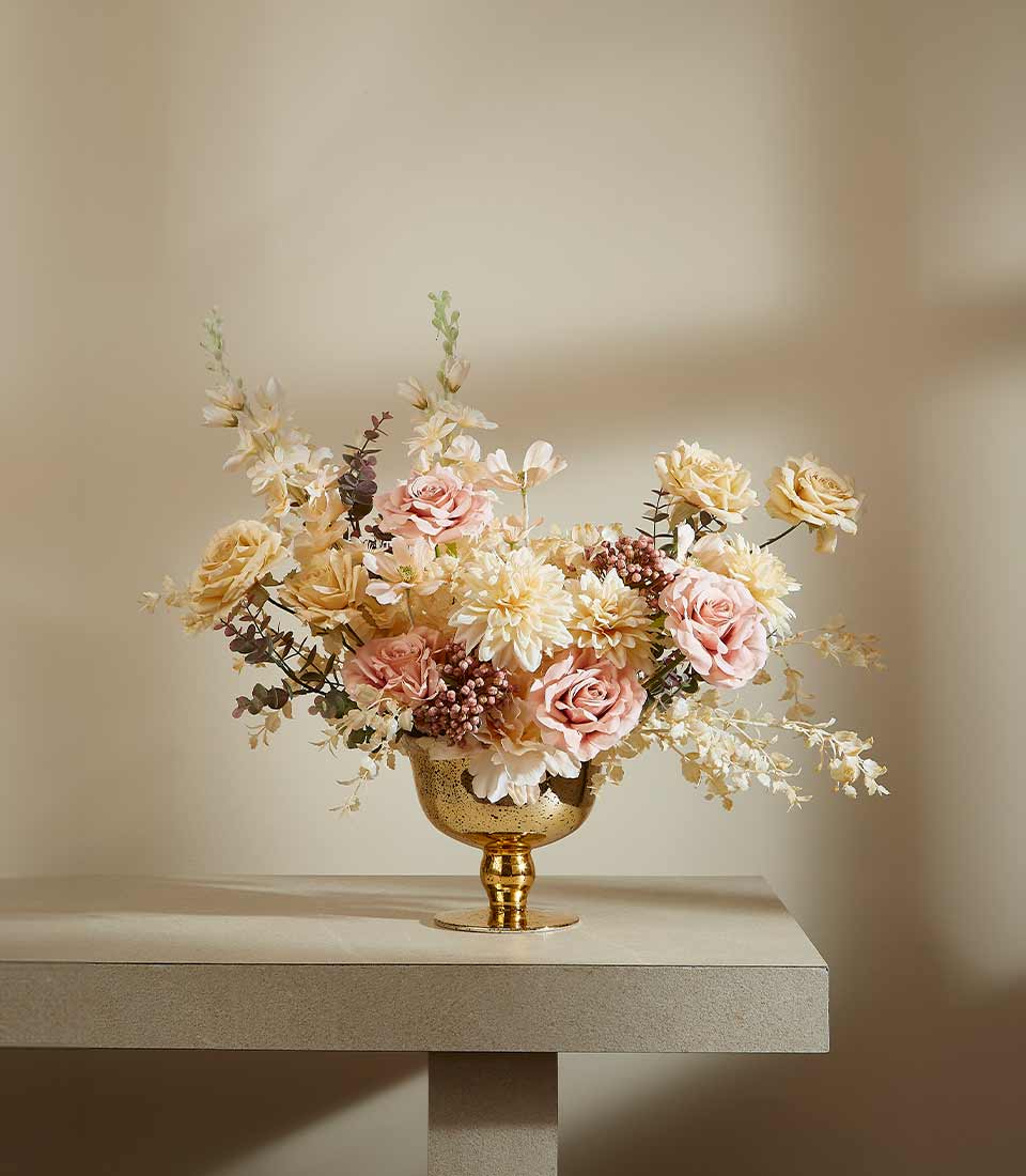 Gold rose artificial flower arrangement in textured metal compote vase with soft pink, cream, and pale yellow blooms