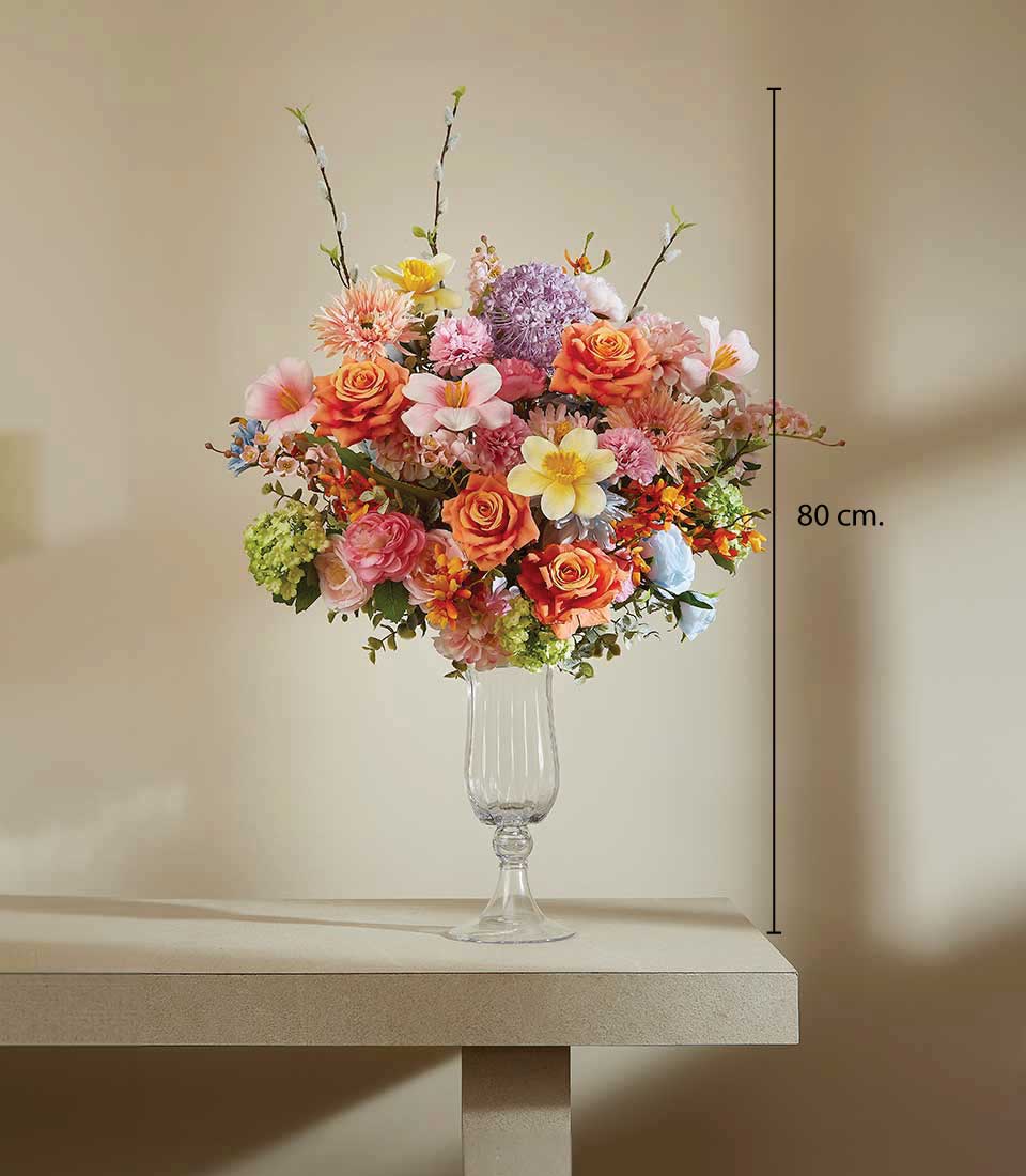 Vibrant artificial flower arrangement in clear glass vase with roses, hydrangeas, and mixed blooms in orange, pink, yellow,