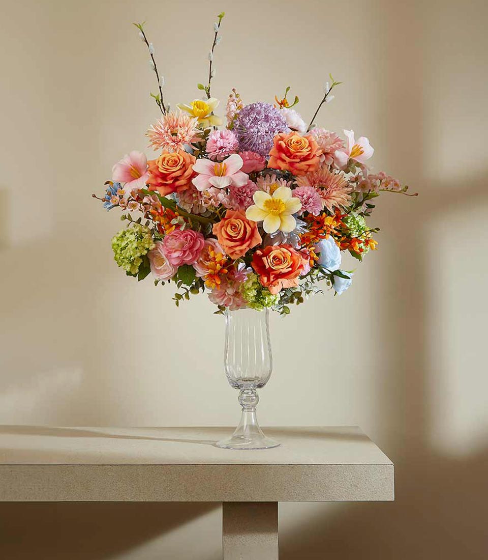 Vibrant mixed artificial flower arrangement with orange roses, pink lilies, and purple alliums in fluted glass pedestal vase