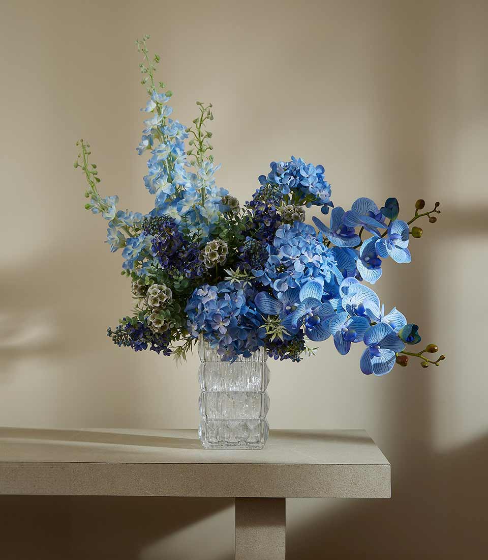 Blue artificial flower arrangement with delphiniums, hydrangeas, and orchids in textured glass vase