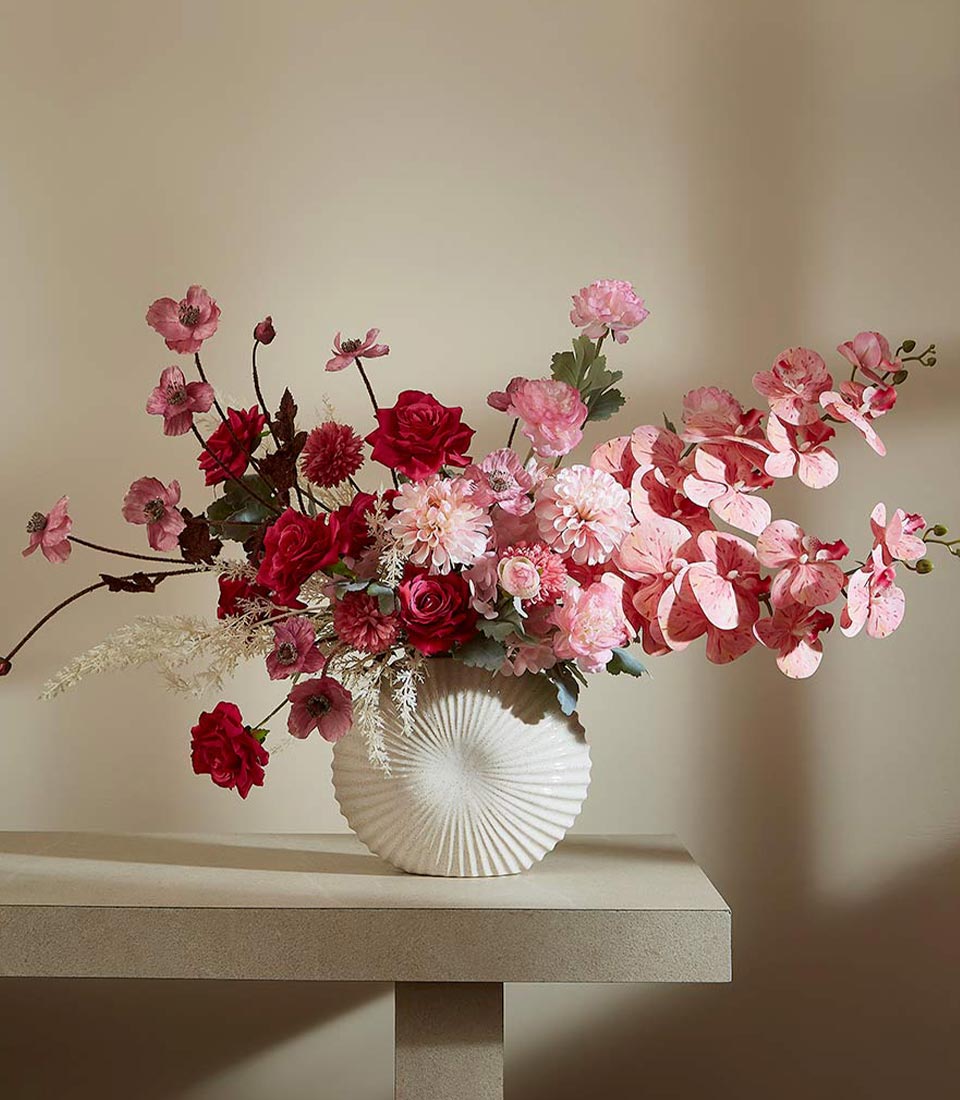 Premium artificial flower arrangement in textured white ceramic vase with roses, dahlias, and orchids in deep red and blush
