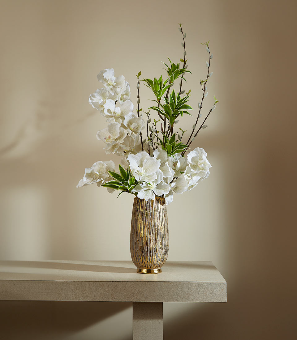 Tall white orchids with green foliage in textured gold ridged vase, elegant artificial flower arrangement