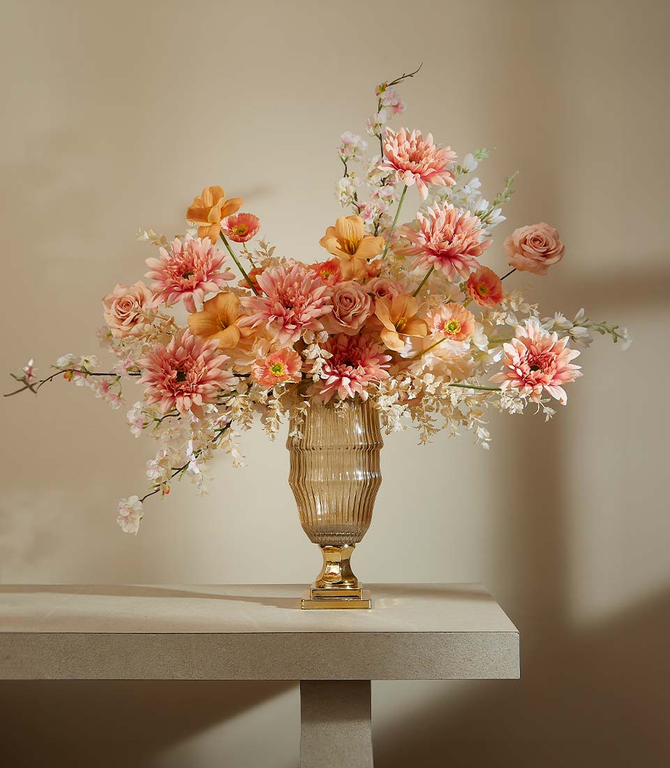 Tall amber glass vase with peach, coral, and orange artificial dahlias and roses on gold pedestal base