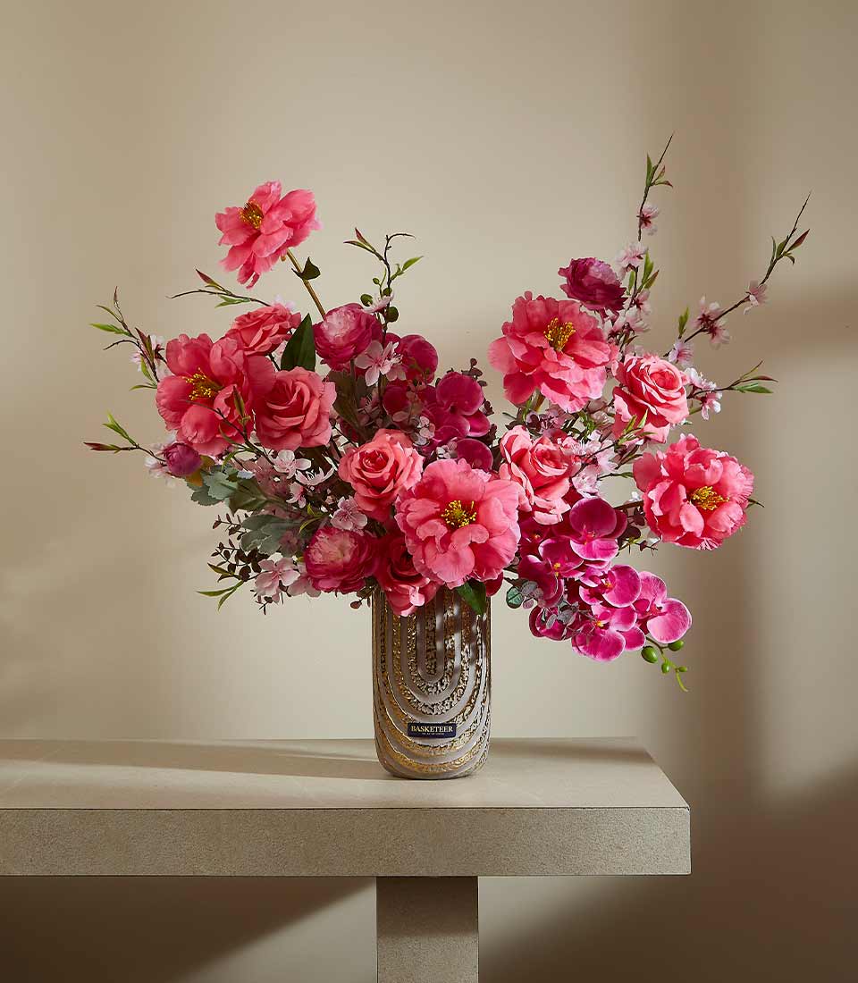 Pink and fuchsia artificial flower arrangement with peonies and roses in gold-patterned glass vase