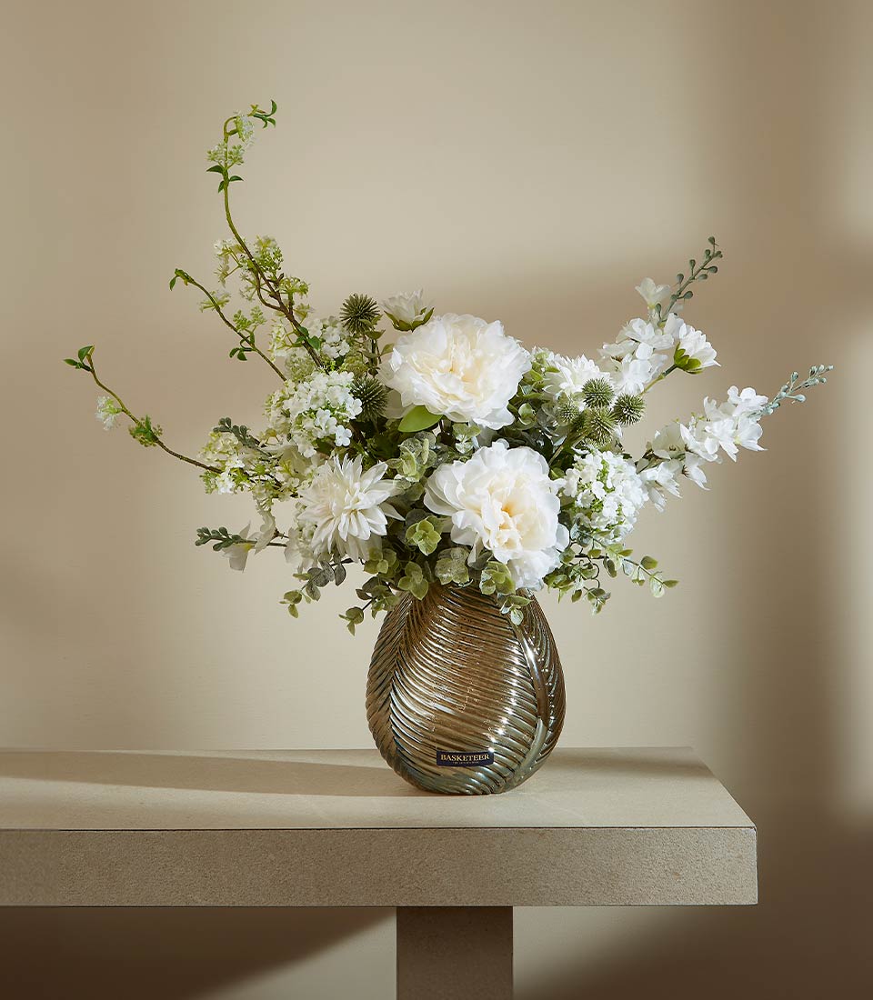 Elegant white peony and dahlia arrangement in bronze-toned ribbed glass vase with green foliage and thistles