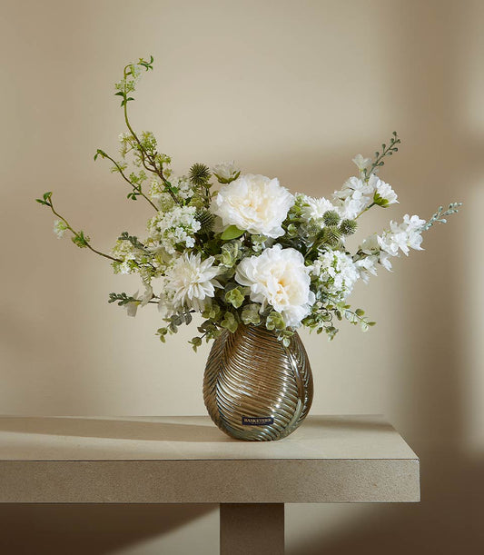 Elegant white peony and dahlia arrangement in bronze-toned ribbed glass vase with green foliage and thistles