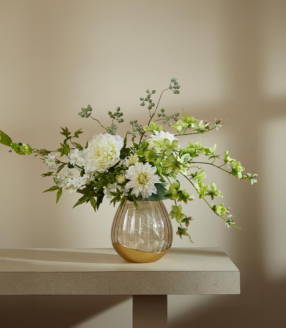 Amber ribbed glass vase with gold base holding white peonies, dahlias, and green foliage artificial flower arrangement
