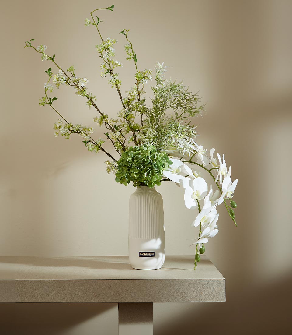 Tall white ceramic vase with artificial white blossoms, green hydrangea, and cascading orchids in modern floral arrangement