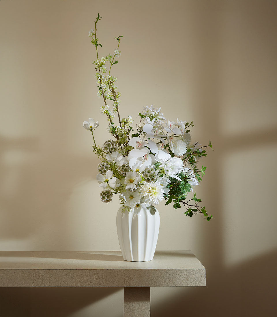 White ceramic ribbed vase with artificial white orchids, dahlias, and green foliage arrangement