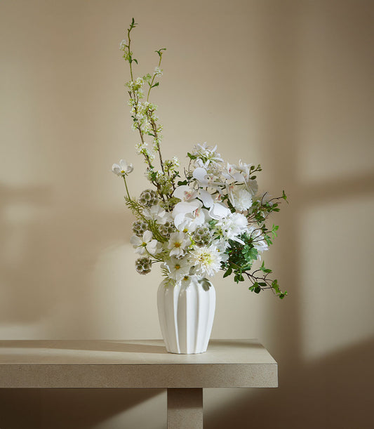 White ceramic ribbed vase with artificial white orchids, dahlias, and green foliage arrangement