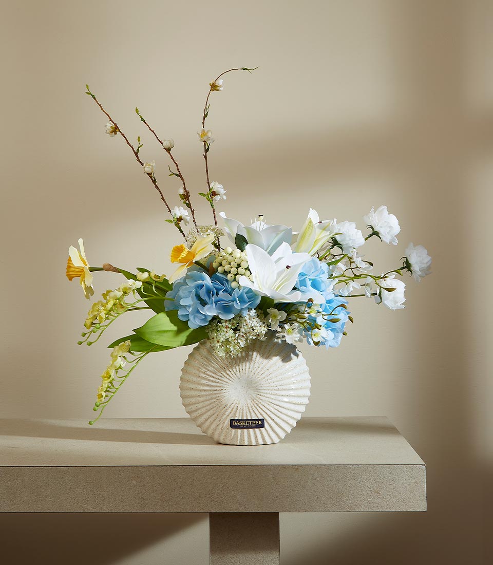 Spring bloom artificial flower arrangement in textured white ceramic vase with lilies, hydrangeas, and daffodils