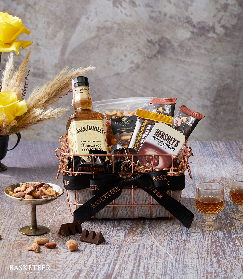 Rose gold wire gift basket with Jack Daniel's Honey whiskey, chocolates, nuts, and black ribbon on rustic wood