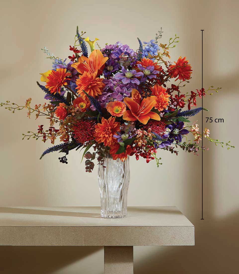 Vibrant artificial flower arrangement in purple, orange, and red with lilies and hydrangeas in textured glass vase
